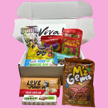 Open treat box with various vegan goodies- sweets and chocolate on a pink background