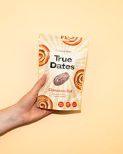 A hand presents a white-and-yellow True Dates package labeled “Cinnamon Roll,” decorated with cinnamon-roll swirls against a warm peach background.