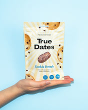 A hand holds a cream-and-yellow True Dates Cookie Dough pouch against a bright blue background