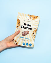 A hand holds a cream-and-yellow True Dates Cookie Dough pouch against a bright blue background