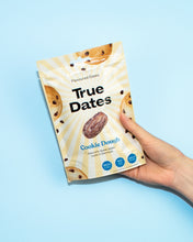 A hand holds a cream-and-yellow True Dates Cookie Dough pouch against a bright blue background