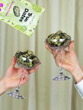 Two hands hold stemmed glasses filled with dark, green-speckled date bites beneath a True Dates package, creating a playful, celebratory mood.