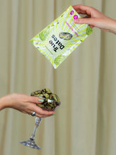 A hand pours a green-and-white dates pouch toward a stemmed glass holding scoops of dark, chunky dessert; the playful setup feels casual and appetizing.