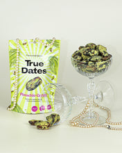 A pistachio-cream True Dates package stands beside a glass bowl piled with dates; spilled dates and looping pearl strands create an elegant, celebratory still life.