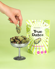 A hand lifts a pistachio-coated date above a bowl of dates beside a bright green True Dates Pistachio Cream pouch, creating a fresh, playful display.