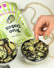 A manicured hand picks a green-coated pistachio flavoured dates from a silver bowl beside a True Dates snack pouch and looping pearl necklace, styled as elegant, playful indulgence.