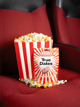 A red-and-white striped popcorn tub and True Dates caramel popcorn package sit on a plush red cinema seat, evoking a cozy movie-night treat.