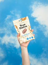 A hand holds a cream-colored True Dates Cookie Dough snack pouch against a vivid blue, cloud-dappled sky, creating a bright, upbeat feel.
