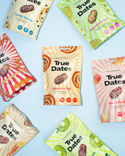 Colorful True Dates snack packages arranged on a pale blue background, featuring date varieties like Caramel Popcorn, Cinnamon Roll, and Pistachio Cream; the playful, bright design feels cheerful and lively.