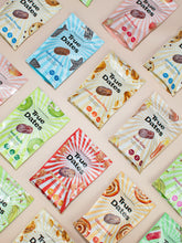 Angled view of colourful True Dates snack packages scattered on a pale background, featuring date illustrations and bold patterns in blue, pink, green, cream, and red; bright, playful tone.