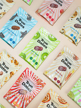 Angled view of colourful True Dates snack packages scattered on a pale background, featuring date illustrations and bold patterns in blue, pink, green, cream, and red; bright, playful tone.