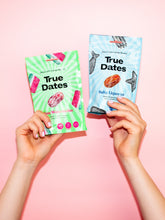Hands hold two True Dates snack pouches against a pastel pink background: green watermelon and blue salty licorice flavors, in a bright, playful product display.