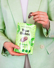 A person in a mint-green suit holds a bright green True Dates Sour Apple snack pouch and a chocolate-covered date, presenting the product against a pale background.