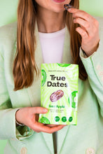 A person in a mint-green suit holds a bright green True Dates Sour Apple snack pouch and a chocolate-covered date, presenting the product against a pale background.
