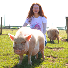A smiling woman with long red hair stands outdoors behind a pink pig, wearing a white graphic T-shirt with pink text and illustration. Another pig is visible in the background on grassy ground.