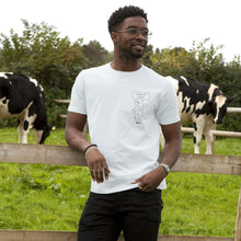 A man wearing a white graphic t-shirt stands in a grassy field with cows behind him. The t-shirt features a pig pencil drawing line art graphic of a pig with a speech bubble that reads 'Free all Pigs'.