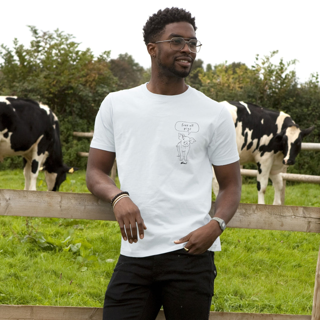 A man wearing a white graphic t-shirt stands in a grassy field with cows behind him. The t-shirt features a pig pencil drawing line art graphic of a pig with a speech bubble that reads 'Free all Pigs'.