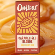 Red Ombar Caramelised Blonde creamy white chocolate bar wrapper, featuring a golden cacao pod and sunburst; labeled organic, natural, fair trade. Background is caramel 