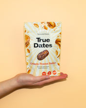 A hand holds a cream-and-yellow True Dates pouch labeled “Creamy Peanut Butter,” with date and peanut images against a warm yellow background. 