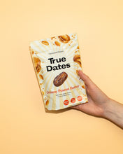 A hand holds a cream-and-yellow True Dates pouch labeled “Creamy Peanut Butter,” with date and peanut images against a warm yellow background. 