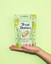 A hand presents a green-and-white pouch of True Dates Pistachio Cream, featuring a date illustration, against a bright lime-green background. The image feels fresh and upbeat.