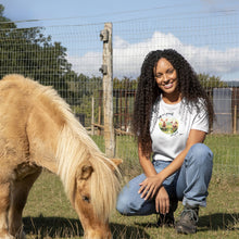 A smiling woman with long curly dark hair crouches outdoors on grass beside a small tan pony, wearing a white graphic T-shirt, which has a design on saying 'pigs are friends not food' and a watercolour of two piglets. A wire fence and trees are visible in the background.
