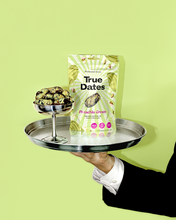 A gloved server holds a silver tray with a bowl of dates and a packet of True Dates Pistachio Cream against a bright lime-green background.