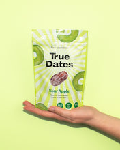 A hand holds a bright green-and-white True Dates Sour Apple flavoured dates pouch against a lime background, with apple and date images; fresh, playful tone.