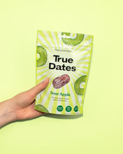 A hand holds a bright green-and-white True Dates Sour Apple flavoured dates pouch against a lime background, with apple and date images; fresh, playful tone.