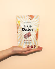 A hand holds a cream-colored pouch of True Dates Sour Cola flavoured dates against a peach background; date illustrations and sunburst rays create a bright, playful feel.
