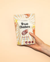 A hand holds a cream-colored pouch of True Dates Sour Cola flavoured dates against a peach background; date illustrations and sunburst rays create a bright, playful feel.