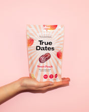 A hand holds a peach-pink pouch of True Dates Sweet Peach against a solid pink background; fruit illustrations and sunburst stripes create a playful, upbeat look.