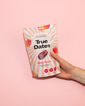 A hand holds a peach-pink pouch of True Dates Sweet Peach against a solid pink background; fruit illustrations and sunburst stripes create a playful, upbeat look.