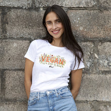 A smiling woman with long dark hair stands against a rustic stone wall wearing a white graphic t-shirt and light blue denim jeans. The t-shirt features a colorful floral design with the text 'VEGAN is a state of kind' and 'Viva!'.