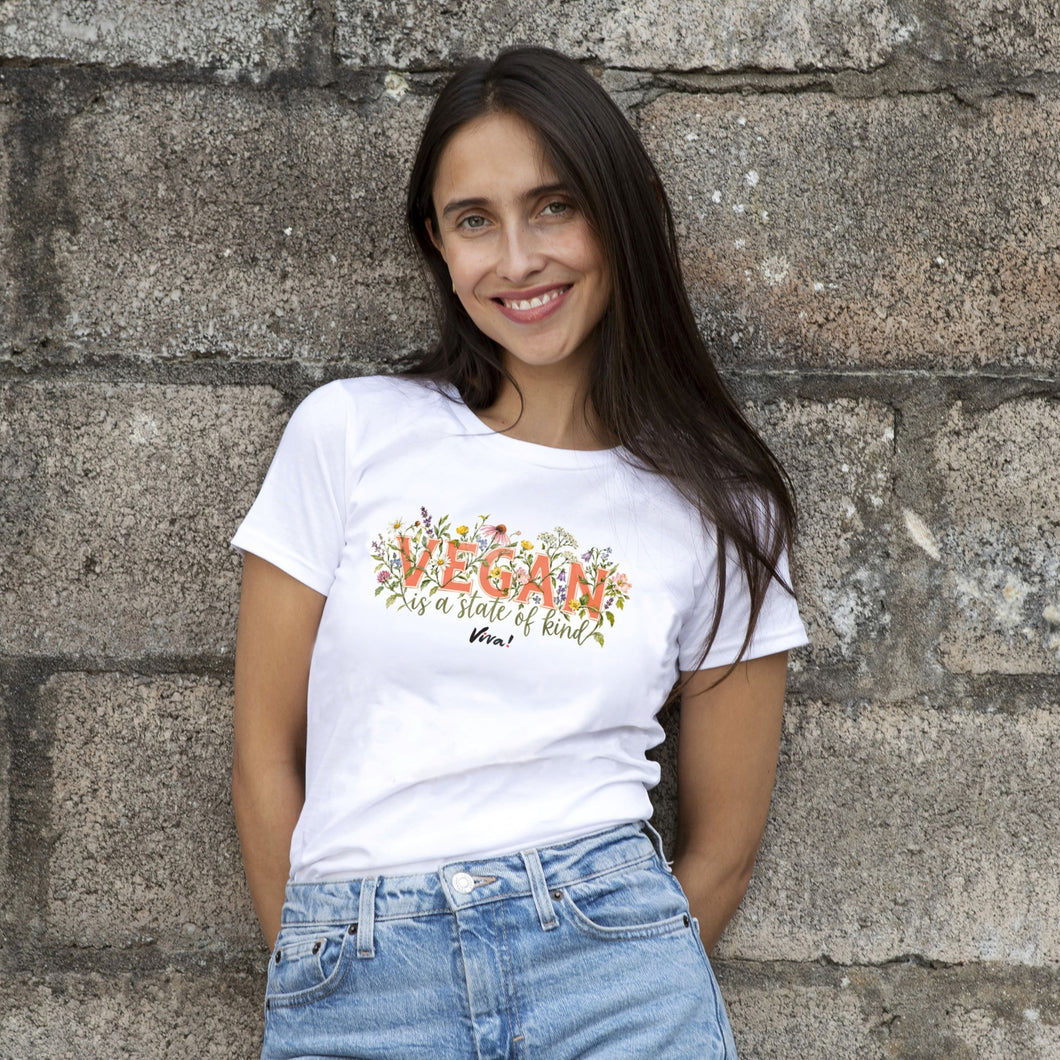 A smiling woman with long dark hair stands against a rustic stone wall wearing a white graphic t-shirt and light blue denim jeans. The t-shirt features a colorful floral design with the text 'VEGAN is a state of kind' and 'Viva!'.